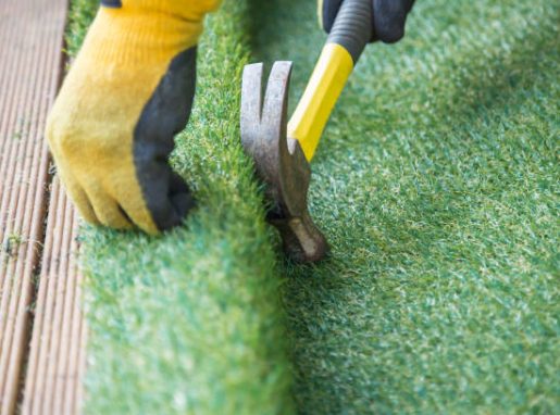 Artificial grass, turf installation alongside decking. A hammer in being used to nail the turf into place. A yellow work glove can be seen.
