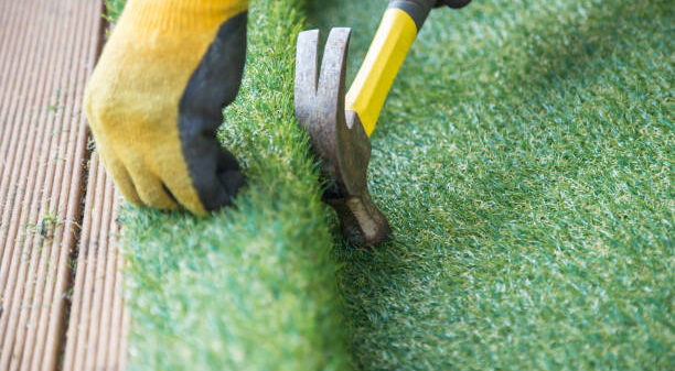 Artificial grass, turf installation alongside decking. A hammer in being used to nail the turf into place. A yellow work glove can be seen.